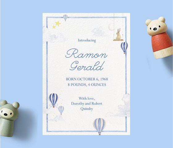 A birth announcement card with light blue text and illustrations of small blue and white hot air balloons, over a light blue backdrop with wooden toys.