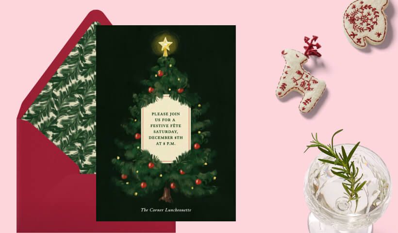 An invitation to a Christmas party with a green, decorated tree set on a black background sits on a red envelope with a holly liner, on a light pink background with knitted ornaments and a cocktail in a clear glass. 