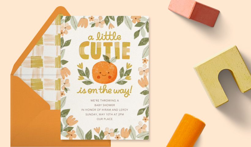 A white invitation with an orange floral border and an orange with a smiley face reading ‘A LITTLE CUTIE IS ON THE WAY!’ on an orange backdrop with wooden blocks. 