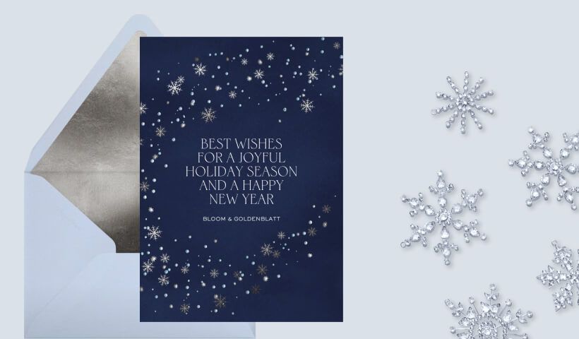 A navy blue card with white snowflakes wishing a joyful season and happy new year, set on a light blue envelope with silver liner, on a grey-blue background with sparkly snowflake ornaments. 