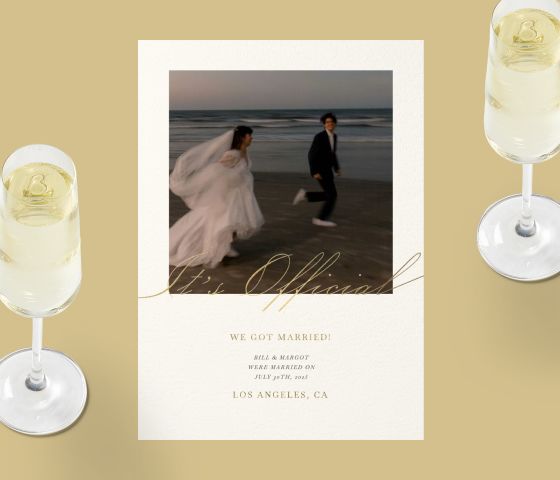 A wedding announcement card with a photo of the bride and groom running on a beach with gold cursive text, over a gold backdrop with two glasses of Champagne. 